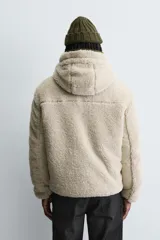Cazadora de corte relajado confeccionada en tejido de textura borreguillo color beige. Presenta cuello subido con capucha ajustable, cierre frontal de cremallera en contraste verde, mangas largas y bolsillos laterales. El interior está forrado en tejido técnico.