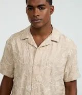 Camisa de poliéster color beige con bordado floral blanco, de corte regular, cuello inglés, manga corta y abotonada en el frente.