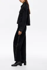 Pantalón de corte barrel color negro, con tiro medio, cierre frontal con botón y cremallera, cinco bolsillos y detalle de tachas metálicas a lo largo de las costuras laterales.