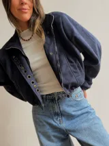 Chaqueta corta de efecto gamuza en color azul marino, con cierre frontal y solapa decorada con una hilera de aros metálicos. Presenta mangas tipo globo, ajuste lateral en la cintura y forro interior.