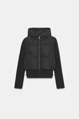 Chaqueta acolchada negra con capucha ajustable, cuerpo con aislamiento térmico y mangas largas en tejido técnico efecto neopreno. Presenta cierre frontal con cremallera, bolsillos laterales y bajo ajustable con cordones elásticos.