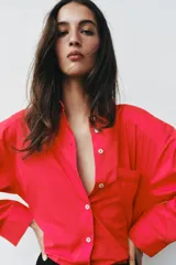 Camisa de popelina roja, de corte oversize, con cuello solapa, manga larga y bajo asimétrico redondeado. Cierre frontal oculto con botones.