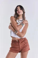 Short corto de tiro bajo confeccionado en gamuza elastizada color marrón camel. Presenta detalles de tachas metálicas doradas en los bolsillos delanteros y en los bordes inferiores, además de una faja en la cintura con hebillas metálicas laterales.