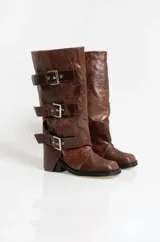 Botas de caña alta confeccionadas en cuero con efecto gastado. Presentan un diseño con tres correas horizontales ajustables mediante hebillas metálicas plateadas. Cuentan con un taco cuadrado bajo y suela de goma.