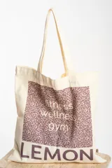 Bolsa tipo tote bag color crudo, con tiras de hombro, confeccionada en 100% algodón. Presenta un diseño gráfico frontal con estampado en tonos morados que incluye texto con las palabras "fitness, wellness, gym" y la marca "LEMON" en la parte inferior.