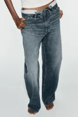 Jeans de mezclilla de tiro medio, corte wide leg y diseño relaxed. Presenta detalles rotos y desgastados en las piernas, y una cinturilla doblada superpuesta con trabillas y cierre frontal con cremallera y botón metálico.