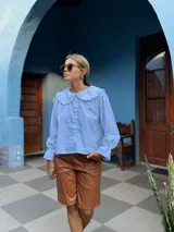 Camisa blanca de corte holgado con cuello bebé con volados, cierre frontal de botones y mangas largas con puños terminados en volados.