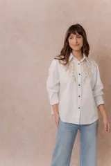Camisa de denim azul con bordados florales en tonos amarillos y celestes sobre los hombros y el pecho. Presenta cuello clásico, cierre frontal con botones y mangas largas con puños.