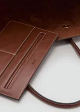 Bolso tipo tote marrón de gran tamaño, con diseño estructurado, dos asas superiores y detalles de hebillas en los laterales. Cuenta con forro interior y cierre de botón magnético.