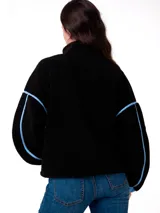 Buzo polar de textura suave en color negro, con cierre frontal parcial y detalles en color celeste en el cuello, puños y costuras laterales. Presenta un corte holgado y el logo de la marca bordado en el pecho.