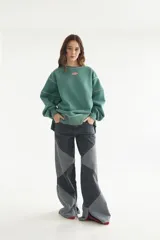 Buzo verde de algodón con diseño holgado y cuello redondo. Cuenta con costuras decorativas expuestas en color claro y un pequeño parche bordado en el pecho.
