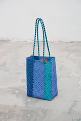 Bolso tipo matera tejido artesanalmente con tiras de plástico en tonos degradados de azul, violeta y turquesa, con diseño geométrico y correas trenzadas.