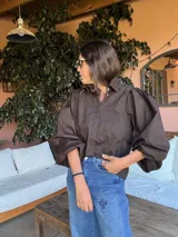 Camisa de color marrón oscuro con cuello clásico y mangas abullonadas con puños elásticos. Presenta un corte holgado y cierre frontal con botones.