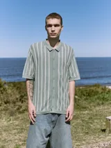 Polo de punto verde con rayas verticales blancas, corte relajado, cuello tipo camisa, cierre con botones y mangas cortas.