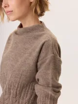 Buzo de lana color beige con cuello chimenea, mangas largas y diseño de tejido trenzado en la parte inferior y puños.