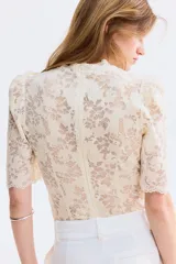 Top de encaje color crema con ribetes festoneados, cuello de volados, mangas cortas abullonadas, alforzas en el frente y cierre oculto en la parte posterior.