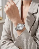 Reloj analógico de pulsera con caja redonda de metal plateado, bisel decorado con cristales y malla de eslabones metálicos. Cuenta con movimiento de cuarzo japonés y cierre desplegable con botón de seguridad.