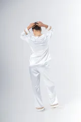 Pijama blanco de satén elastizado, compuesto por una camisa de manga larga con escote en V, abotonada adelante y con puntilla en escote y puños, y un pantalón recto con bolsillos laterales, elástico en la cintura y puntilla en el ruedo.