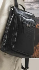Mochila de diseño rectangular con textura símil cuero efecto cocodrilo en color tostado. Cuenta con un bolsillo frontal con cierre, compartimento principal con doble cierre, correas ajustables y manija superior. Incluye detalles metálicos y placa con logo de la marca.