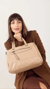 Cartera de mano y hombro color beige, de material símil cuero, con forma estructurada y redondeada. Posee dos asas cortas superiores y una correa larga ajustable y desmontable. Cuenta con un bolsillo exterior con cierre en la parte frontal.