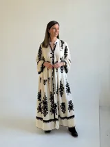 Vestido largo color marfil con estampado ornamental en color negro, escote en V con botones frontales, mangas largas amplias y diseño escalonado en la falda.