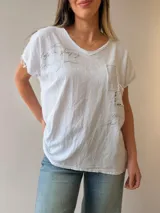 Remera de lino y algodón color beige con escote en V, mangas cortas y calce relajado. Presenta un bolsillo frontal decorado con apliques de strass, encaje y tachas, además de sutiles estampas con escritura manuscrita y detalles desflecados.