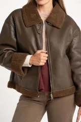 Campera tipo gamulán de cuero sintético en color marrón, con interior y detalles en cuello y puños de corderito sintético al tono. Presenta un corte oversize con cierre frontal metálico.