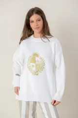 Buzo de tejido de punto color blanco, de corte holgado y cuello redondo. Presenta un bordado central en hilo dorado con el escudo nacional y un pequeño detalle bordado en la manga derecha con los colores de la bandera uruguaya.