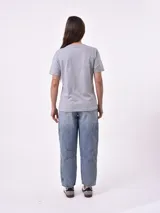 Remera de manga corta color beige claro con cuello redondo. Presenta un estampado gráfico central con un plato de pasta rodeado por un borde azul y blanco, sobre un fondo de franjas verticales rojas y blancas. El estampado incluye texto en cursiva en tonos turquesa y rojo.