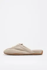 Zapatilla estilo mula de color beige, confeccionada en una combinación de algodón y serraje vacuno, con suela de caucho con tachuelas texturizadas y costuras decorativas visibles en la parte superior.