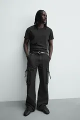 Pantalón cargo de corte relajado en color negro con efecto lavado. Presenta bolsillos laterales tipo cargo con solapas y cintas ajustables, bolsillos frontales y traseros, y detalles de argollas metálicas. Cierre frontal con cremallera y botón.