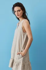 Vestido corto de lino color beige, de silueta suelta y sin mangas, con escote en V decorado con un botón, bolsillos laterales y detalle de volado en el ruedo.