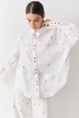 Camisa de lino blanca con estampado de figuras deportivas pequeñas y detalles de costuras en color rojo. Presenta cuello clásico, cierre frontal con botones de madera, mangas largas con puños abotonados y un bolsillo frontal aplicado.
