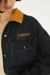 Chaqueta oversize de denim azul oscuro con cuello camisero de pana color mostaza, cierre frontal con botones metálicos y bolsillos delanteros con solapa. Cuenta con detalle de bordado en el pecho.