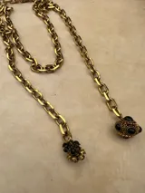 Collar de cadena de eslabones gruesos en tono dorado, con diseño tipo lazo que termina en dos dijes colgantes: uno esférico con detalles labrados y piedras negras, y otro con forma de flor decorado con cristales negros.