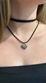 Gargantilla tipo choker compuesta por una cinta negra lisa y un collar largo con dije de corazón de metal plateado con diseño labrado.