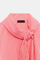 Camisa de rayas rosadas y blancas, con cuello subido decorado con un lazo integrado, manga larga con puños abotonados y cierre frontal oculto.