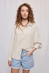 Camisa de manga larga color beige con cuello clásico y botonadura frontal. Presenta mangas con volumen sutil, puños abotonados y una silueta amplia de caída fluida.
