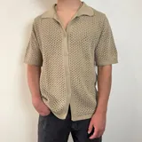 Camisa de tejido de punto calado en color beige, con cuello polo, cierre frontal de botones y mangas cortas.