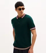 Polo de manga corta en punto texturizado de color verde oscuro, con cuello y puños ribeteados en bordó y blanco con rayas. Cuenta con cierre frontal parcial de botones.