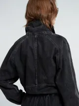Mono de denim negro con cierre frontal de botones, cintura elástica y detalle de flecos en la parte superior delantera. Presenta mangas largas con puños elásticos y corte recto en las piernas.