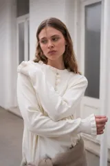 Sweater de punto color crema, con cuello redondo y cierre frontal parcial con botones. Presenta mangas largas con puños acanalados y detalle de volados en los extremos.
