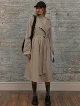 Trench coat de corte midi en color beige, con cuello de solapa amplio, cierre frontal con cinturón ajustable a la cintura, bolsillos laterales y detalle de botones en los puños.