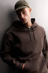 Sudadera de hombre Zara Athleticz de ajuste relajado (relaxed fit), color marrón, confeccionada en tejido técnico compacto con mezcla de algodón y elasticidad. Cuenta con cuello con capucha ajustable con cordones negros, manga larga, bolsillo frontal tipo canguro con un pequeño bolsillo termofijado con cierre, y acabados con rib en puños y cintura.