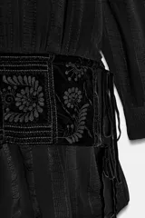 Vestido corto de viscosa color negro con cuello perkins y manga larga. Presenta un fajín de terciopelo integrado con bordados a tono y detalle de lazadas laterales.