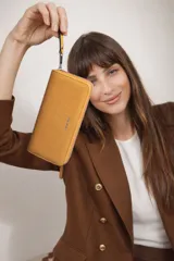 Billetera larga de diseño rectangular con cierre perimetral, confeccionada en material sintético texturizado color mostaza. Presenta el logo de la marca grabado en el frente y una correa de mano corta para mayor comodidad.