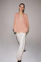 Blusa de viscosa color rosado con cuello redondo, manga larga abullonada con puños elásticos y cierre frontal con lazadas. Presenta un diseño con volantes en el bajo y corte asimétrico.