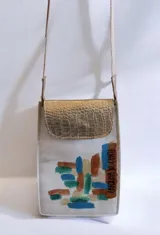 Bandolera compacta de cuero color blanco, con solapa de cuero texturizado metalizado dorado y diseño pintado a mano en el frente. Cuenta con cierre de broche imán y correa larga de cinta textil con detalles en cuero grabado.
