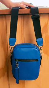 Bandolera morral color beige y blanco, con correa ajustable de tela en color blanco. Tiene cierre superior y un bolsillo frontal con cierre. Incluye un llavero decorativo.