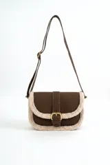 Cartera bandolera marrón de gamuza sintética con detalles de corderito sintético color beige en los bordes. Cuenta con una solapa frontal con cierre de hebilla metálica dorada y correa ajustable.